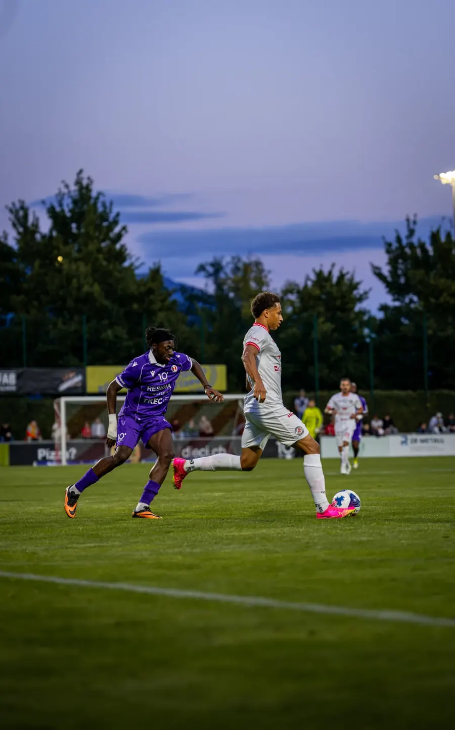FCW-Spieler in Weiss am Ball in der Abenddämmerung, ein Gegenspieler in Lila verfolgt ihn – FC Winterthur gegen FC Rapperswil-Jona, 21. August 2026